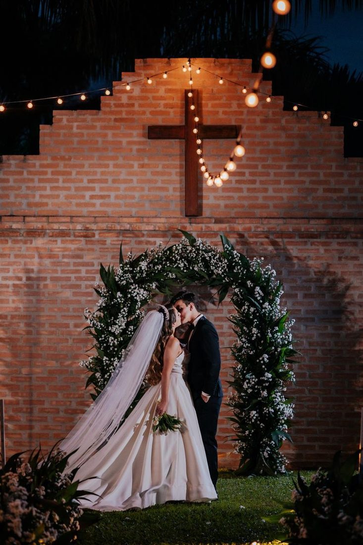 Noivos se beijando sob o arco de flores brancas, diante do muro de tijolos com a cruz de madeira iluminada